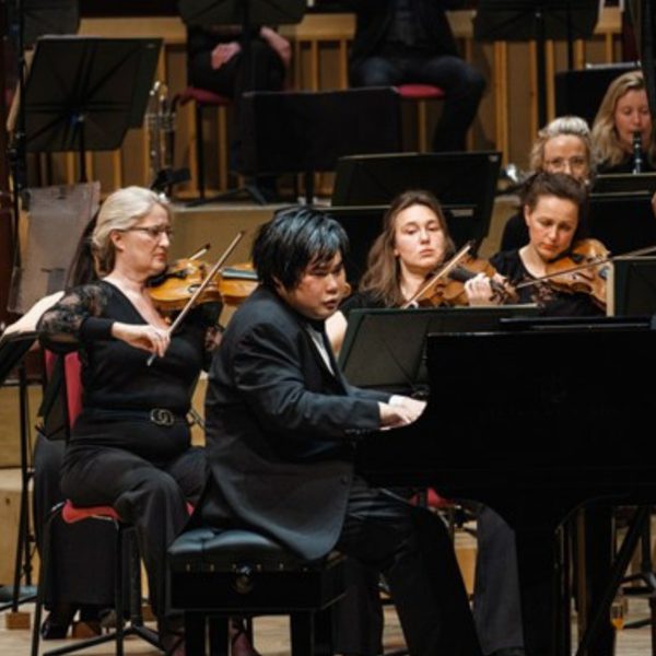 Nobuyuki Tsujii. an Asian man with short black hair, playing the piano on stage surrounded by violinists