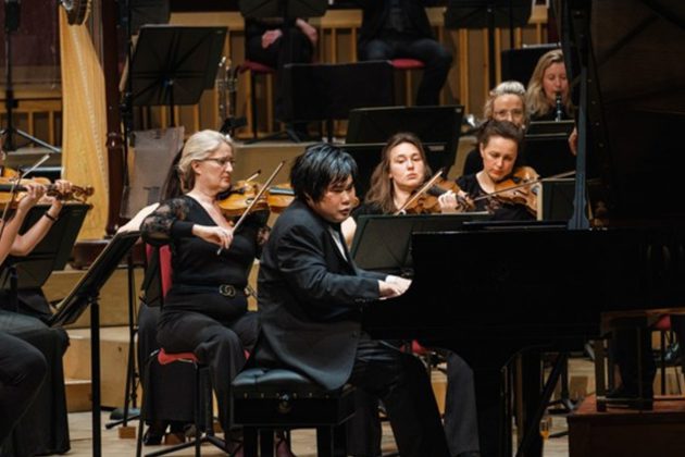 Nobuyuki Tsujii. an Asian man with short black hair, playing the piano on stage surrounded by violinists