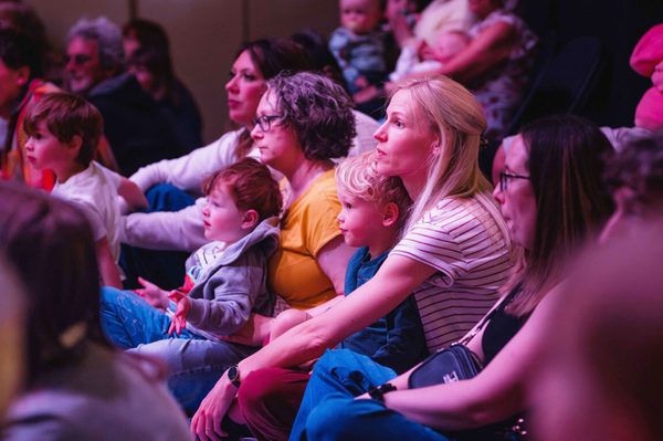 An audience of parents and young children sat on the floor, lit by a pink hue