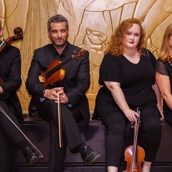 Members of Liverpool String Quartet sat in a line in front of a golden wall, wearing all black and holding their instruments