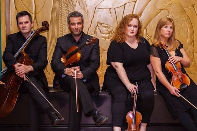 Members of Liverpool String Quartet sat in a line in front of a golden wall, wearing all black and holding their instruments