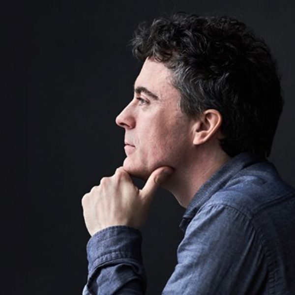 Paul Lewis, a white man with short grey hair, wearing a blue denim shirt and leaning on his hand looking to the left