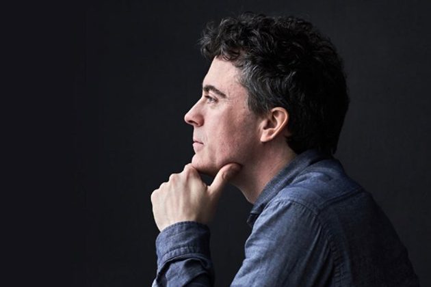 Paul Lewis, a white man with short grey hair, wearing a blue denim shirt and leaning on his hand looking to the left