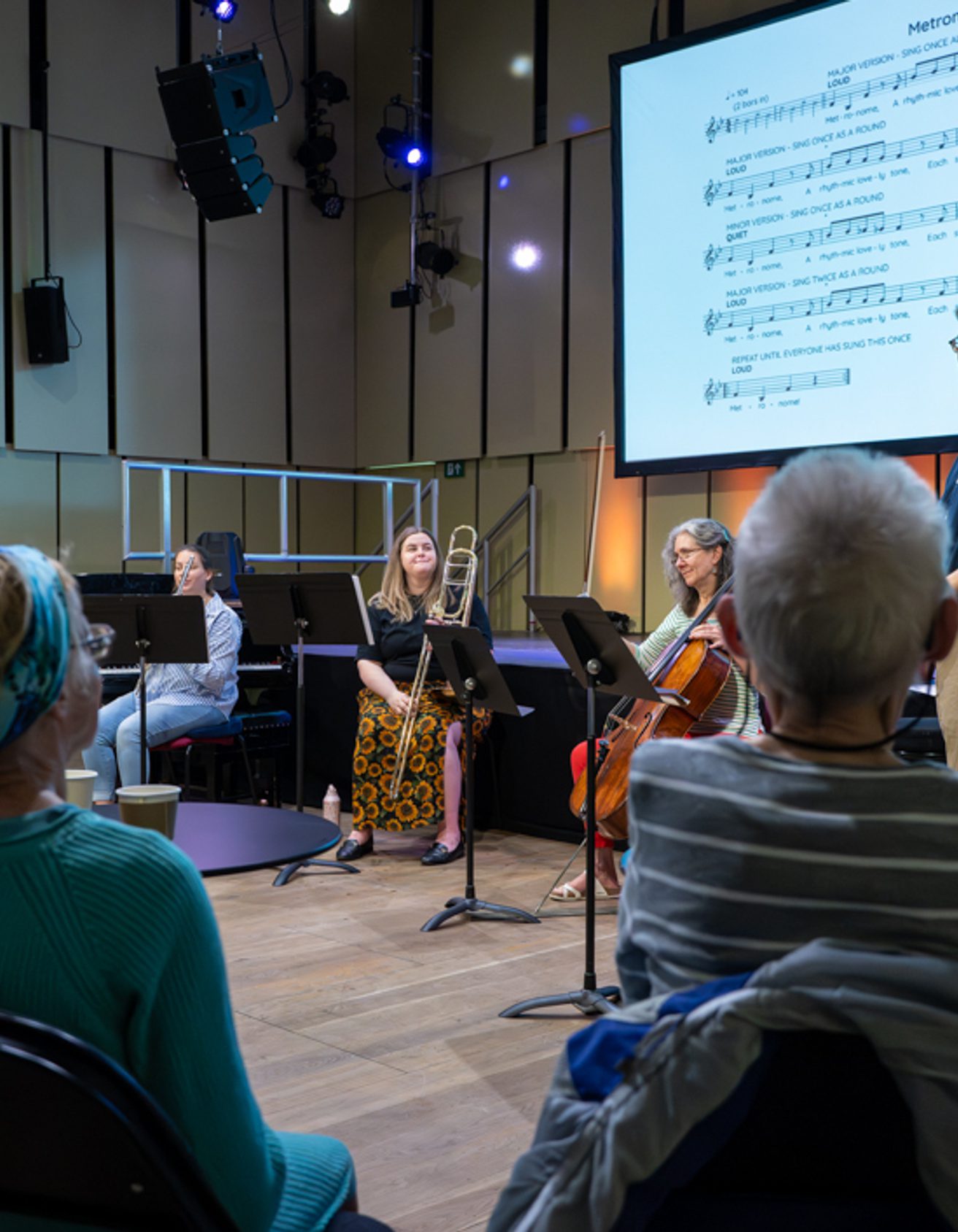 three members of the Orchestra sitting and holding their instruments and Ian is standing next to them wearing a navy blue shirt, talking to a small audience who have their backs to the camera