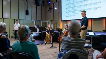 three members of the Orchestra sitting and holding their instruments and Ian is standing next to them wearing a navy blue shirt, talking to a small audience who have their backs to the camera