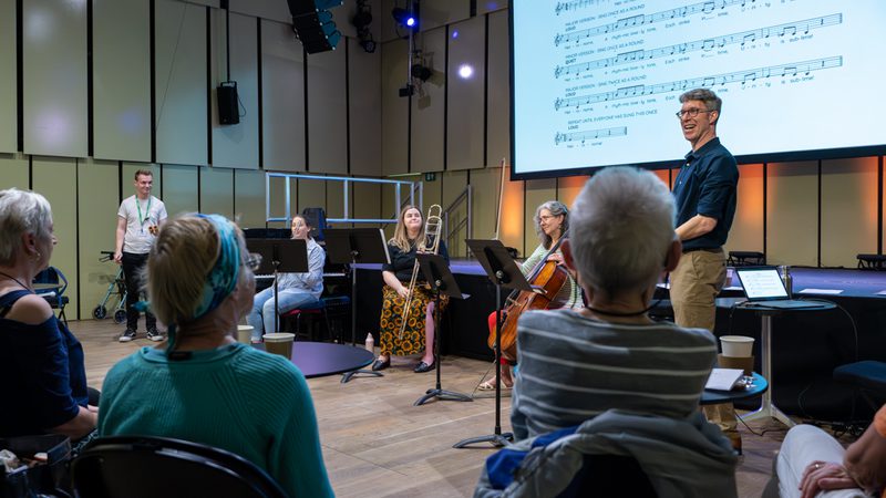 three members of the Orchestra sitting and holding their instruments and Ian is standing next to them wearing a navy blue shirt, talking to a small audience who have their backs to the camera