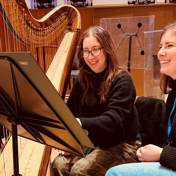 Elizabeth McNulty and Chloe Chousmer-Kerr sitting next to a harp wearing black jumpers smiling at a black music stand