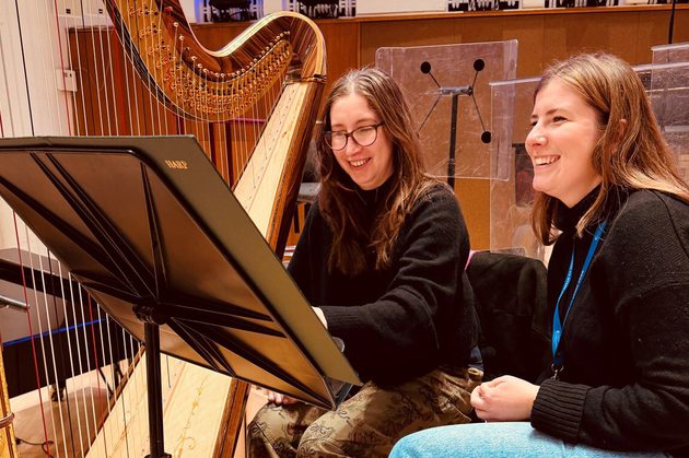 Elizabeth McNulty and Chloe Chousmer-Kerr sitting next to a harp wearing black jumpers smiling at a black music stand
