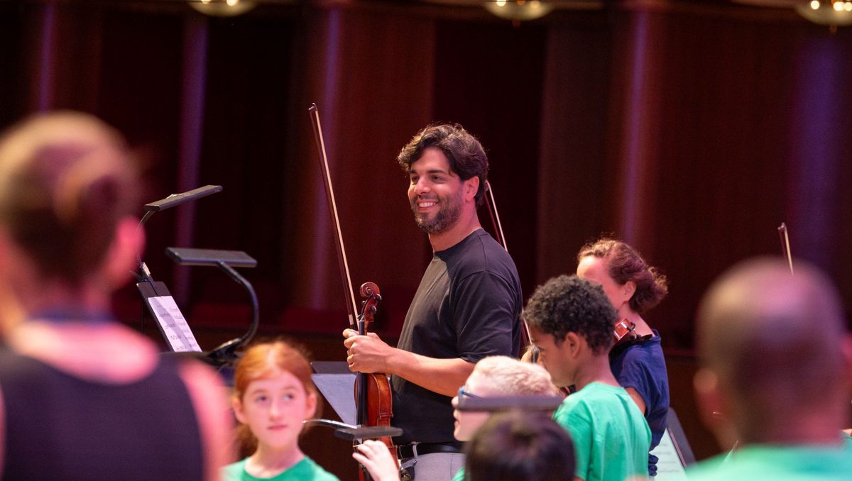Domingo Hindoyan smiling while looking slightly towards the left and holding a violin, some members of In Harmony are standing around Domingo wearing green shirts